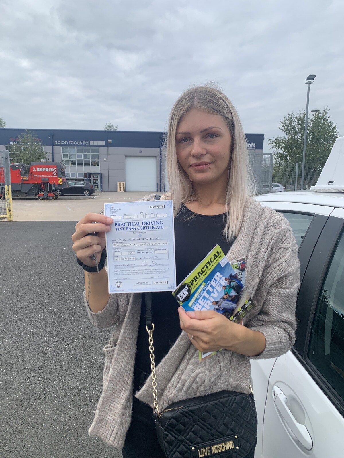 Learner holding a practical driving test pass certificate beside a learner car.