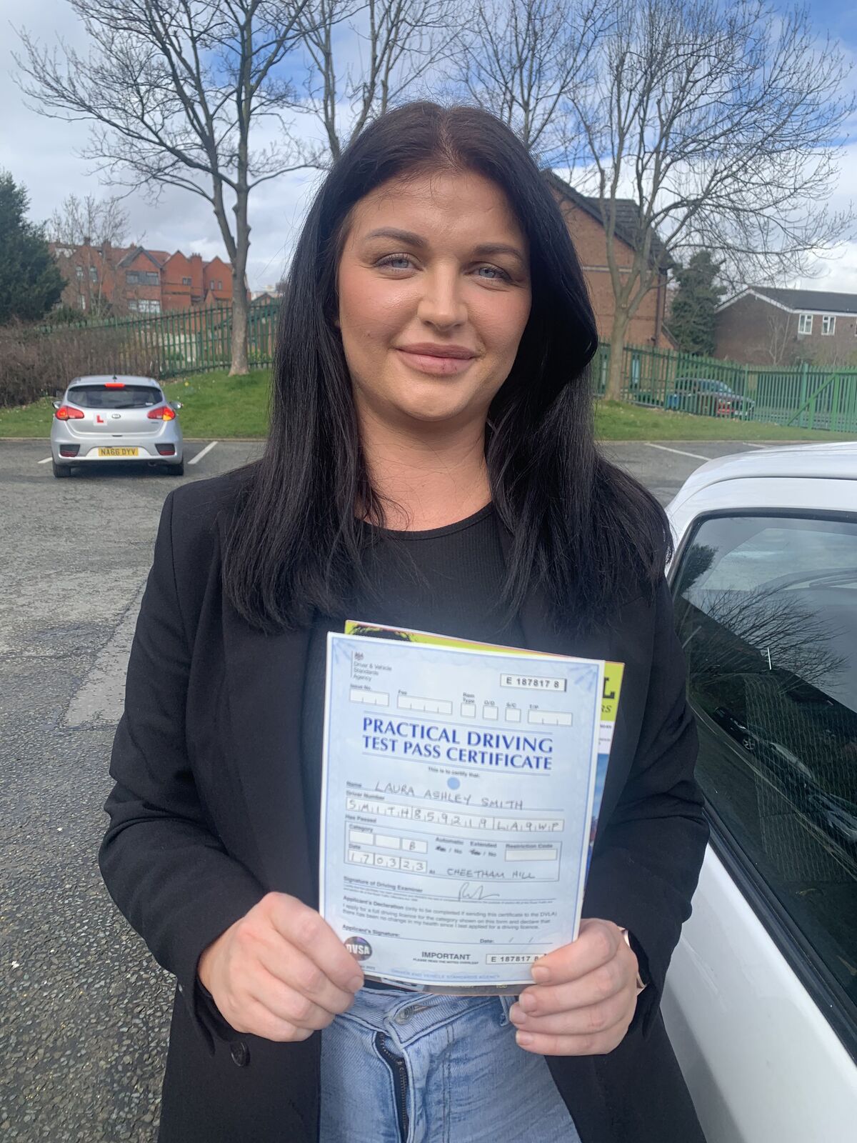 Learner smiling while holding a practical driving test pass certificate beside a car.